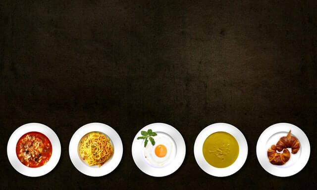 Top view of five plates with different foods including pasta, soup, fried egg, and croissants on a dark background.