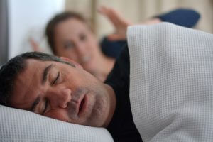 Man sleeping with mouth open while woman in the background looks disturbed in bed.
