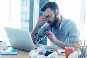 Tired man sitting at desk with laptop, holding his head and glasses, showing signs of stress or fatigue.