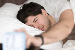 Young man lying in bed, squinting and reaching to turn off the alarm clock in the morning.