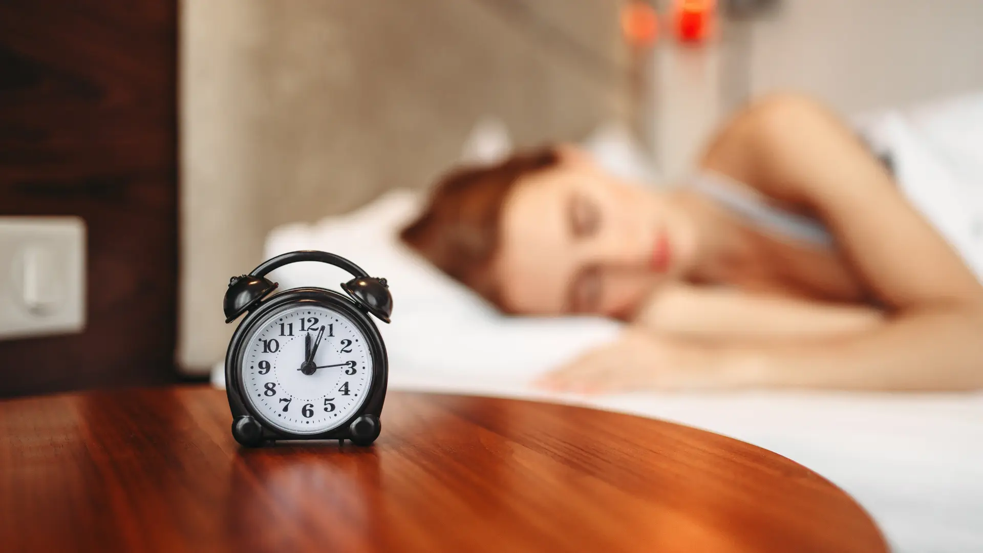 Black alarm clock on a wooden surface with a sleeping woman in the background.