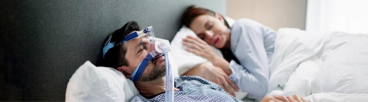 Man wearing CPAP mask for sleep apnea treatment while partner sleeps beside him.
