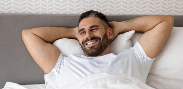Smiling man lying in bed with hands behind head looking relaxed.