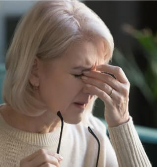 Older woman rubbing her eyes and pinching the bridge of her nose while holding glasses, showing signs of fatigue or eye strain.
