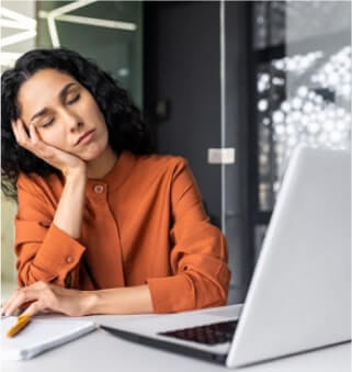 Young woman in orange shirt falling asleep at her desk with laptop open, resting head on hand.