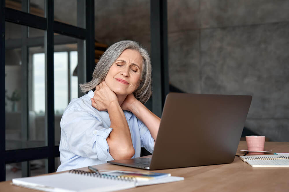 Older woman holding her neck in pain while working on a laptop at a desk
