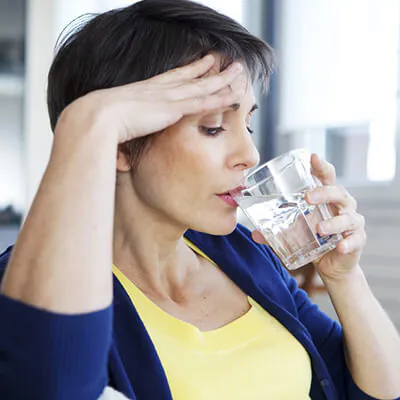 Middle-aged woman holding her forehead while drinking a glass of water, appearing unwell or fatigued