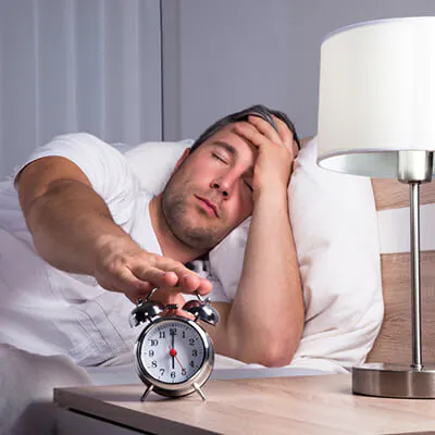 Sleepy man in bed reaching to turn off an alarm clock while holding his head in tiredness.