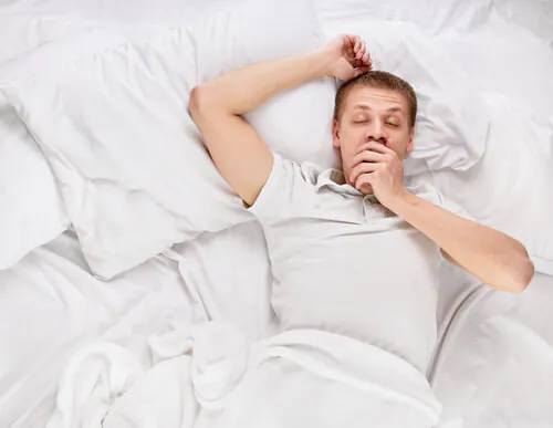 Man lying in bed yawning and looking tired, showing signs of sleepiness.