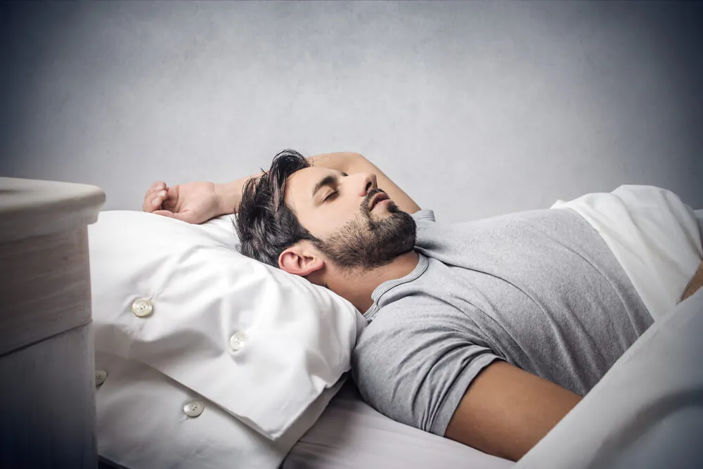 Man sleeping peacefully on his back in bed with one arm resting above his hea