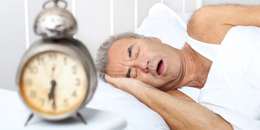 Older man sleeping on his side with mouth open next to an alarm clock on the bedside table