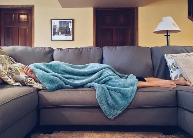 Person lying on a couch covered with a blue blanket, appearing to rest or nap