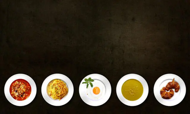 Top view of five plates with different foods including pasta, soup, fried egg, and croissants on a dark background.