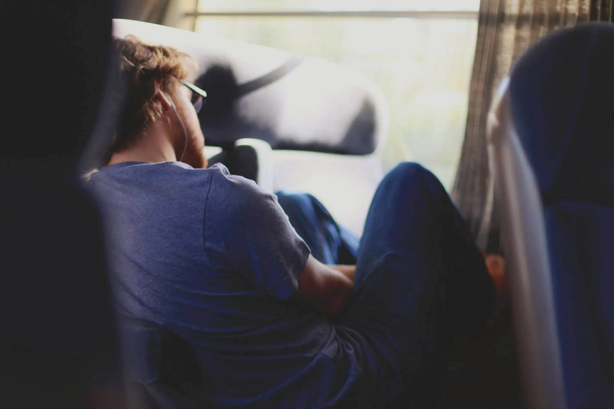 Man sitting by a bus or train window, leaning back with earphones on, appearing to rest or sleep.