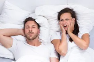 Man sleeping while snoring in bed as a frustrated woman covers her ears beside him