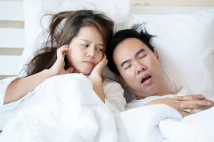 Man snoring loudly in bed while woman beside him covers her ears in frustration.