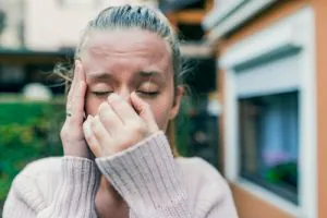 Young woman outdoors holding her nose and forehead, appearing to suffer from sinus pressure or headache.