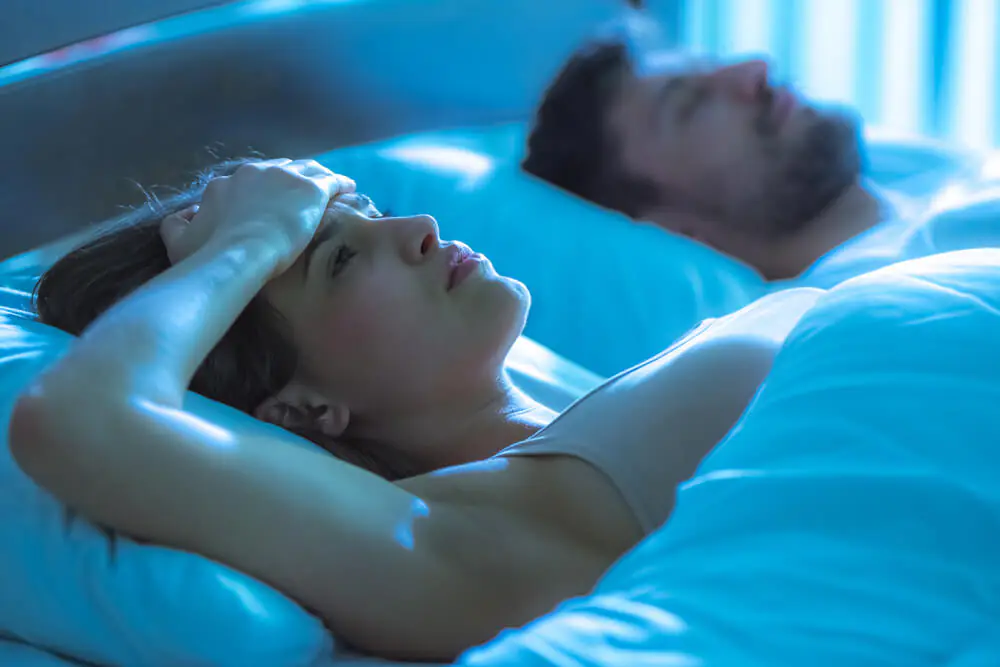 Young woman lying awake in bed with hand on forehead while man sleeps beside her.